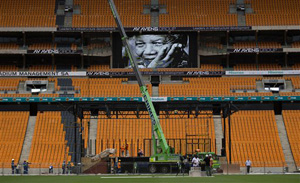 An image of former South Africa President Nelson Mandela is shown on a big screen as work is carried out to put up a stage at the FNB stadium where his memorial service will take place on Tuesday, in Johannesburg, South Africa, Monday, Dec. 9, 2013. Scores of heads of state and government and other foreign dignitaries, including royalty, are beginning to converge on South Africa as the final preparations for Tuesday's national memorial service for liberation struggle icon Nelson Mandela are put in place.