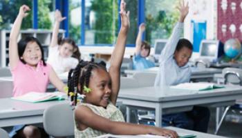 Children in a classroom raising hands to answer the teacher